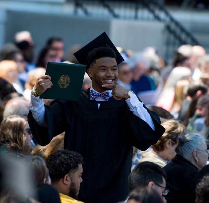 male undergraduate student at commencement pointing to diploma in crowd