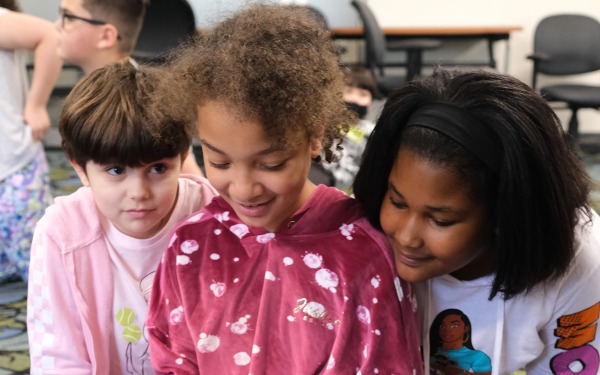 Girl holding caterpillar on the floor in McKay classroom with two other girls looking on