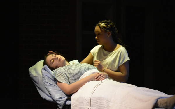 Girl in hospital bed with girl by her side in Stop Kiss Play