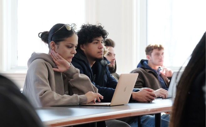 One female on laptop and three male students at table listening to lecture