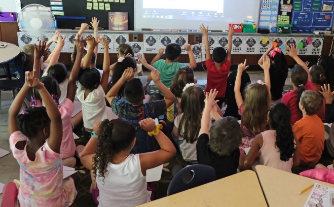 Classroom of students looking at board raising their hands