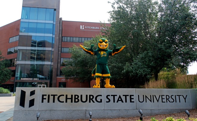 Freddy standing on top of Fitchburg State University sign in front of Hammond.