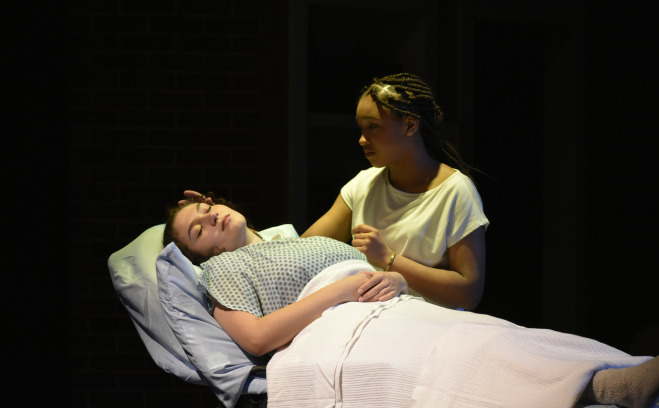 Girl in hospital bed with girl by her side in Stop Kiss Play