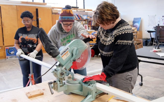 Students and teacher cutting set pieces for play on a saw in shop