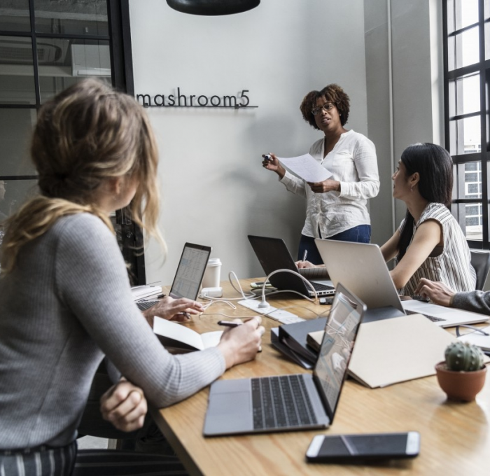 Diverse group of employees sitting around conference table with computers while female presents