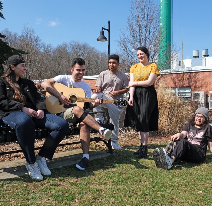 Students on quad sitting on bench playing guitar, singing and laughing