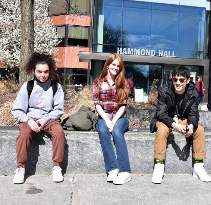 Three students sitting on the wall in front of Hammond Hall on a bright spring day