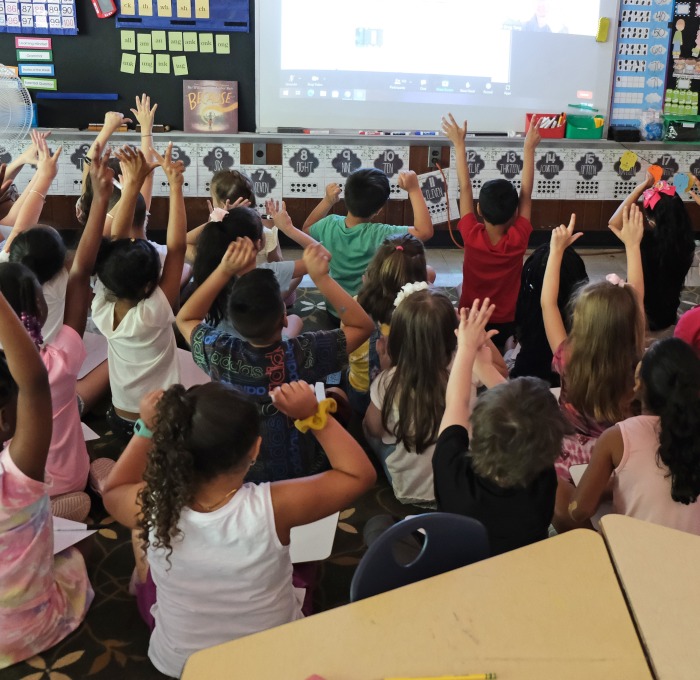 Classroom of students looking at board raising their hands