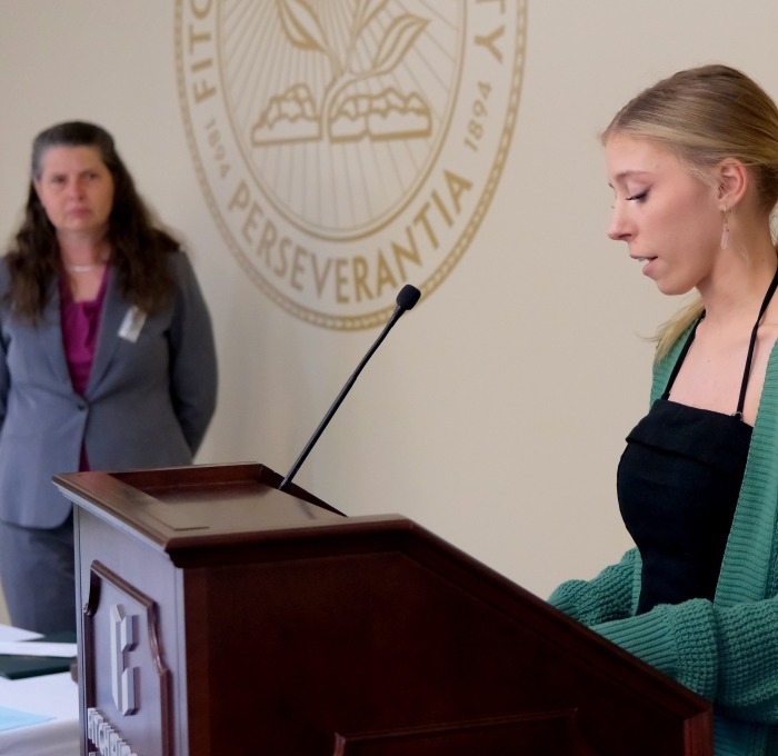 English student giving speech at podium with professor looking on
