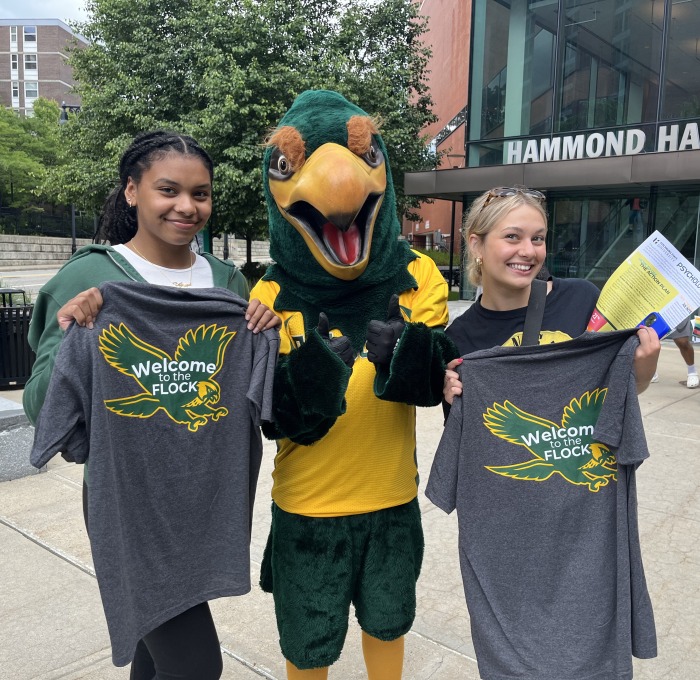 Two female students with Freddy at orientation in front of Hammond with Welcome to Flock shirts