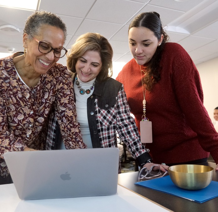 Three teachers in classroom looking at computer with students in background