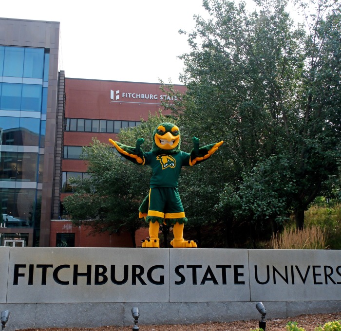 Freddy standing on top of Fitchburg State University sign in front of Hammond.