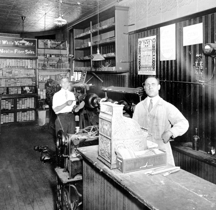 1920 black and white photo of Michele Fraticelli standing behind the cash register of the newly remodeled Cobbler Shop 8 Water Street, Leominster, MA with Mr. Principle with shoe.