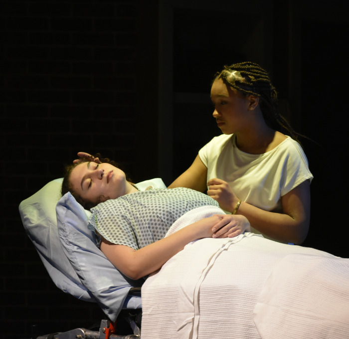 Girl in hospital bed with girl by her side in Stop Kiss Play