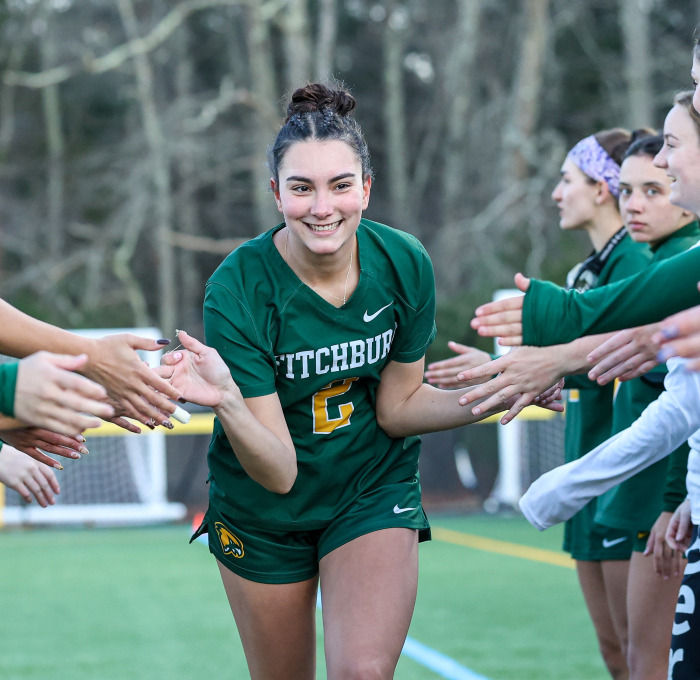 Women's lacrosse player running on the field slapping team mates hands.
