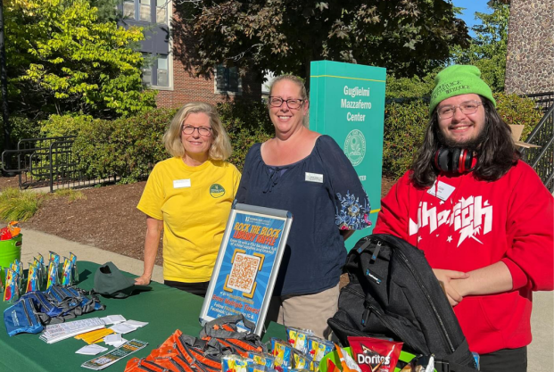Two white women with blond hair and one white man with long brown hair and a bright green knit hat smiling and standing behind a table with snacks and drinks, set up outdoors