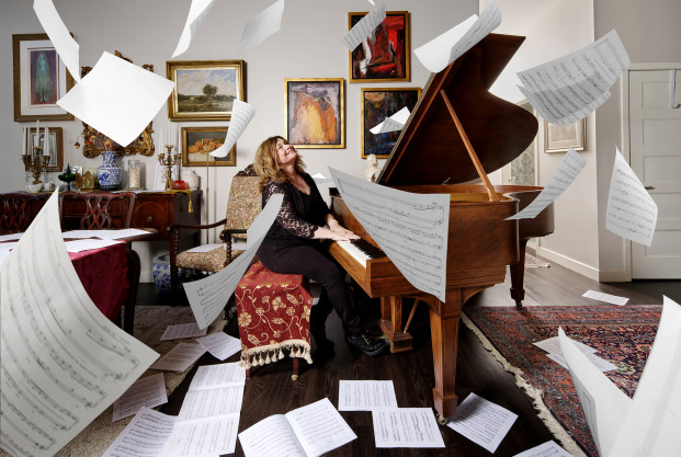 White middle age woman with light brown shoulder length hair sitting at a baby grand piano in an artfully decorated living room looking up towards the ceiling while a swirl of sheet music flutters to the floor 