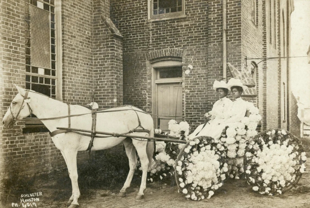 Historic black-and-white photograph featuring two Black women, dressed in elegant white attire and large decorative hats, seated in a floral-adorned horse-drawn cart pulled by a white horse