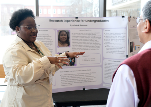 A black woman with short curly hair and glasses wearing beige button blouse is talking to a person in front of a research poster