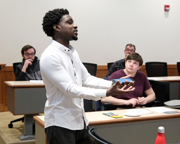 Student in classroom with phone in hand describing project while others look on.
