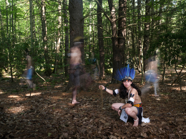 Indigenous teenager with long dark curly hair wearing traditional clothing including a large headdress of bright blue feathers, staring directly into the camera in a squatting position in front of a grove of trees and ghost images of the same boy dancing in a circle