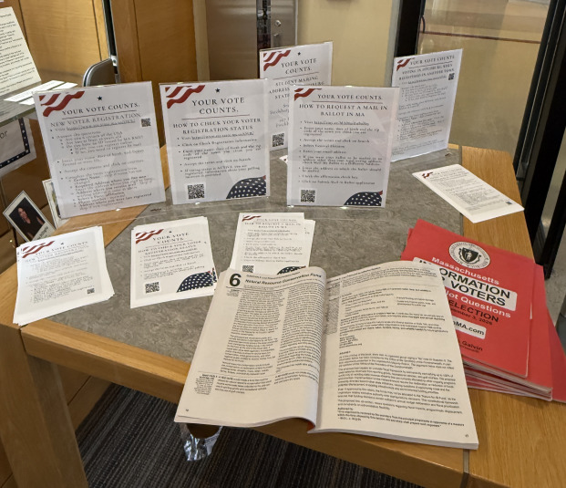 A tabletop display of voter information flyers, QR codes, an open booklet, and Massachusetts election materials on a table.