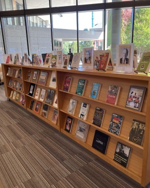 A library display featuring numerous books on wooden shelves, organized neatly. Large windows in the background allow natural light to illuminate the area.