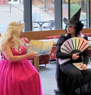 Two drag queen performers in costumes sitting. One is wearing a long blond wig, wearing a bright pick dress and holding a picture book open and reading. The other is looking at her wearing a black witch costume holding a rainbow fan.
