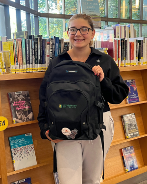 White young woman with glasses and brown hair pulled back, smiling, holding a black backpack posing in front of a bookcase