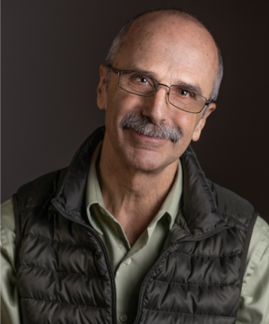 Portrait of Ricardo Barros, white bald man with a gray mustache and glasses looking directly at the camera, wearing a black puffer vest and light green button down shirt