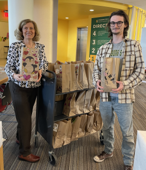 A wooden cart with small paper bags with a white middle age woman with shoulder length blond hair holding a decorated small paper bag standing on the left, and and taller white young man with brown hair and glasses standing on the right holding a small decorated paper bag. 