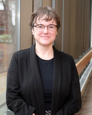 White woman with glasses and short greyish brown hair wearing black shirt and sweater standing in front of a large window