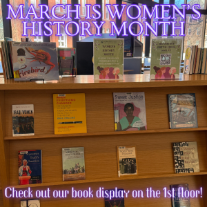 The image shows a library book display celebrating Women's History Month. At the top, large neon-style purple text reads "March is Women's History Month." At the bottom, more neon text says "Check out our book display on the 1st floor!" The background includes windows showing exterior views of buildings.