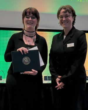 Two people with short dark hair and glasses, smiling. One person is holding a dark folder with a circular emblem.