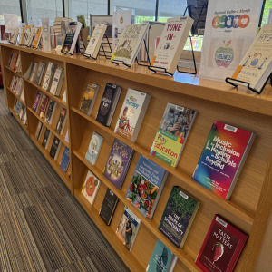 A wooden bookshelf in a library displays a variety of wellness-themed books, with two rows of titles in vibrant colors and illustrations. Sunlight streams through large windows in the background.
