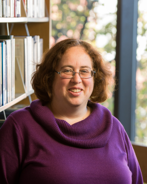 Portrait of a smiling woman with short wavey brown hair wearing glasses and a purple cowl-neck sweater, posed indoors beside a bookshelf and windows.