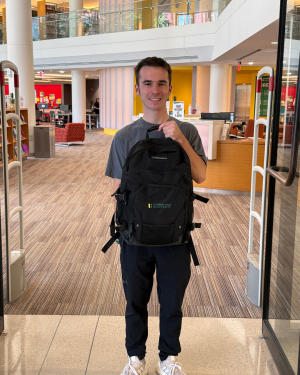 Smiling young white man with short brown hair holding a black Fitchburg State University branded backpack inside the Library entrace